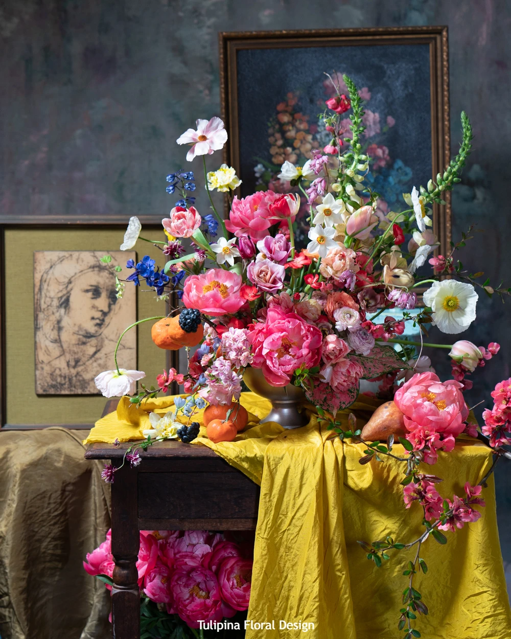 Luxurious Dutch-style floral arrangement featuring coral peonies, pink ranunculus, white anemones, and blue delphiniums displayed on golden velvet cloth with classical artwork backdrop at Holland flower trade show