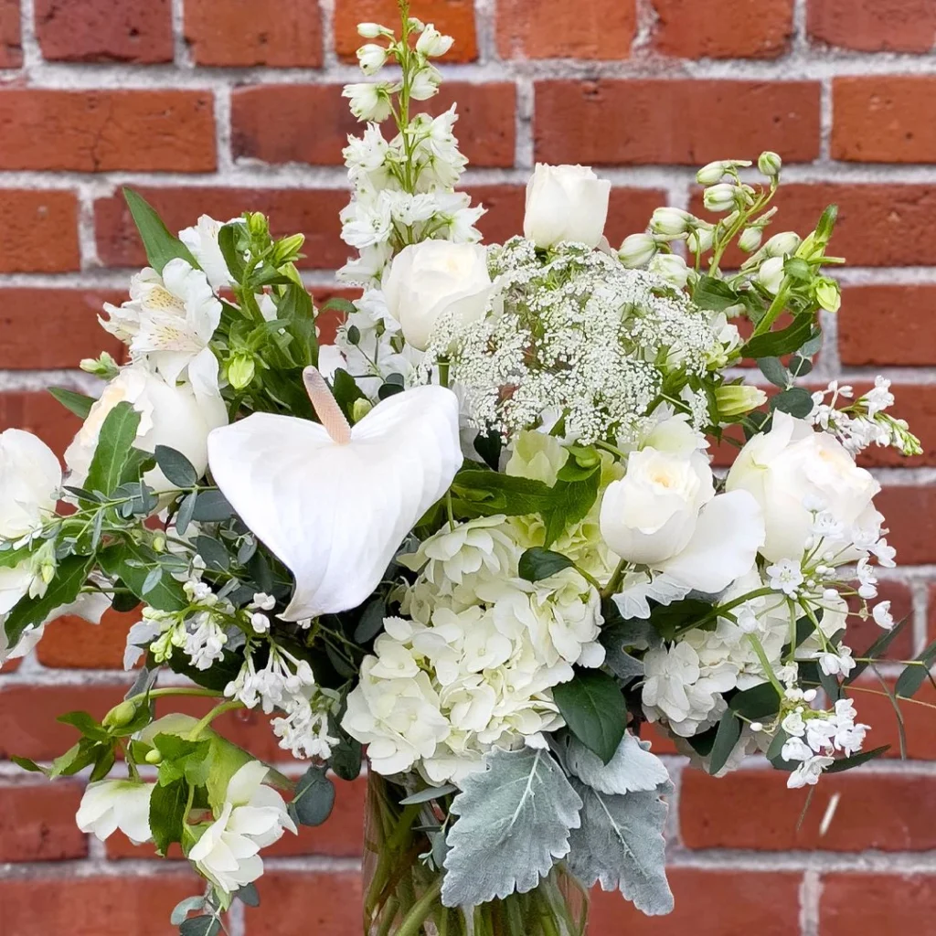 A white wedding-style floral arrangement in a clear vase featuring hydrangea, garden roses, anthurium, queen anne’s lace, eucalyptus, dusty miller, and mixed white accent blooms arranged in loose natural layers in front of a red brick wall.